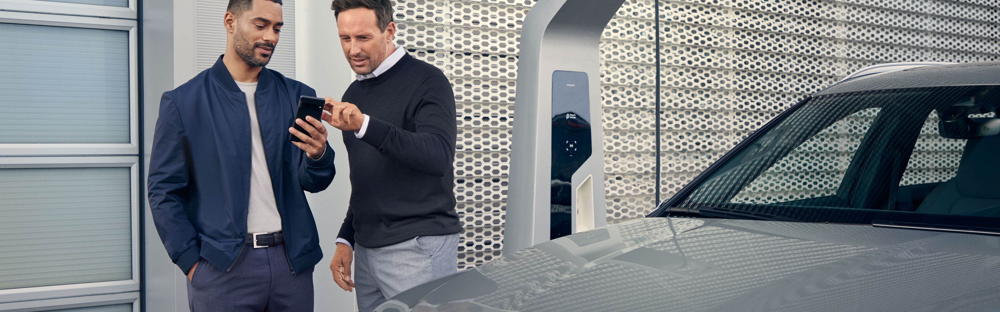 Ein Audi Service Mitarbeiter und ein Kunde blicken auf ein Smartphone, im Vordergrund steht ein Audi Elektroauto