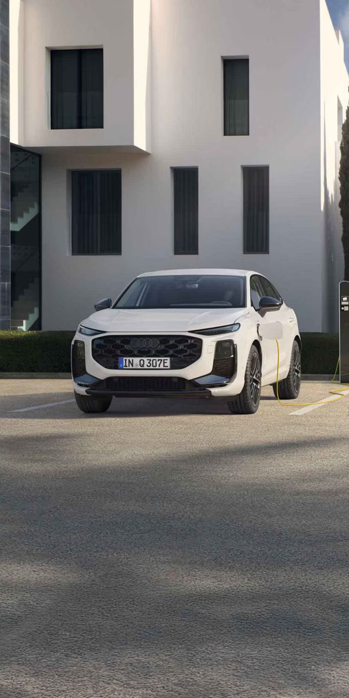 Auf dem Bild ist der Audi Q3 Sportback e-hybrid zu sehen. Dieser steht vor einem Wohngebäude mit einer Ladesäule und lädt.