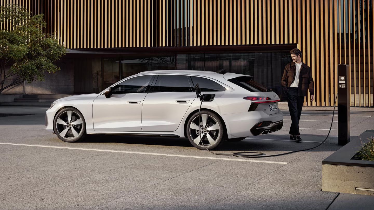 Der A6 Avant e-hybrid steht seitlich vor dem Eingangsbereich eines großen Gebäudes und wird über ein Stromkabel geladen. Am Heck des Fahrzeugs steht ein Mann mittleren Alters.