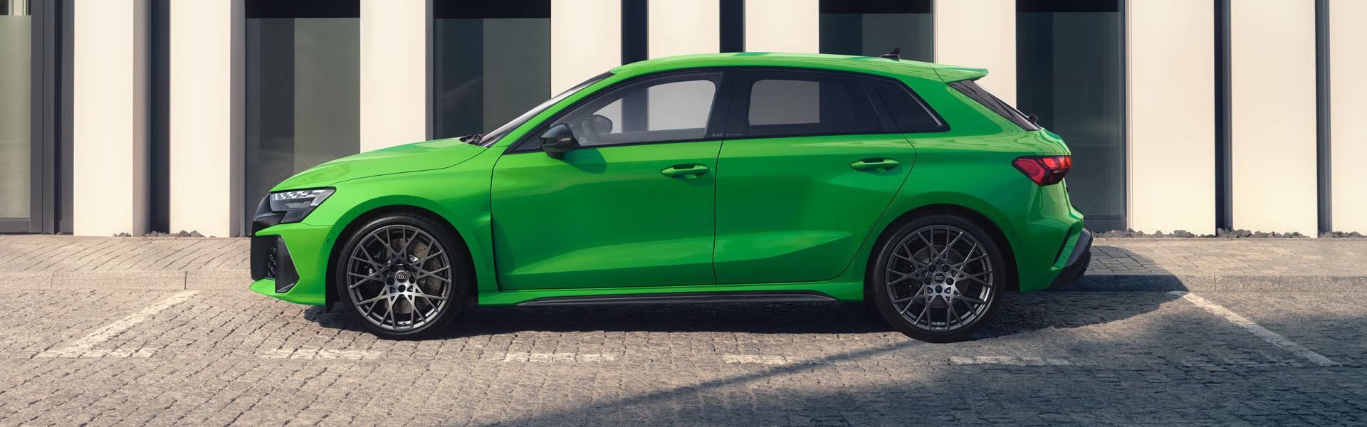 Der RS 3 Sportback von der Seite parkend auf Kopfsteinpflaster, im Hintergrund Umrisse eines modernen Gebäudes.