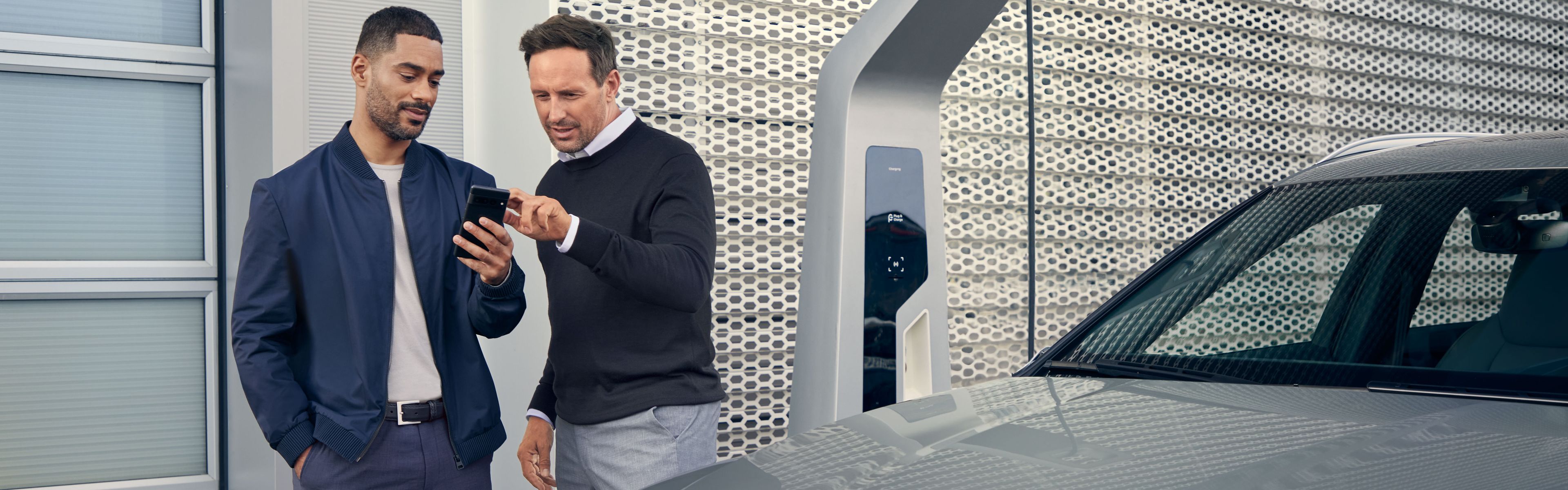 Ein Audi Service Mitarbeiter und ein Kunde blicken auf ein Smartphone, im Vordergrund steht ein Audi Elektroauto