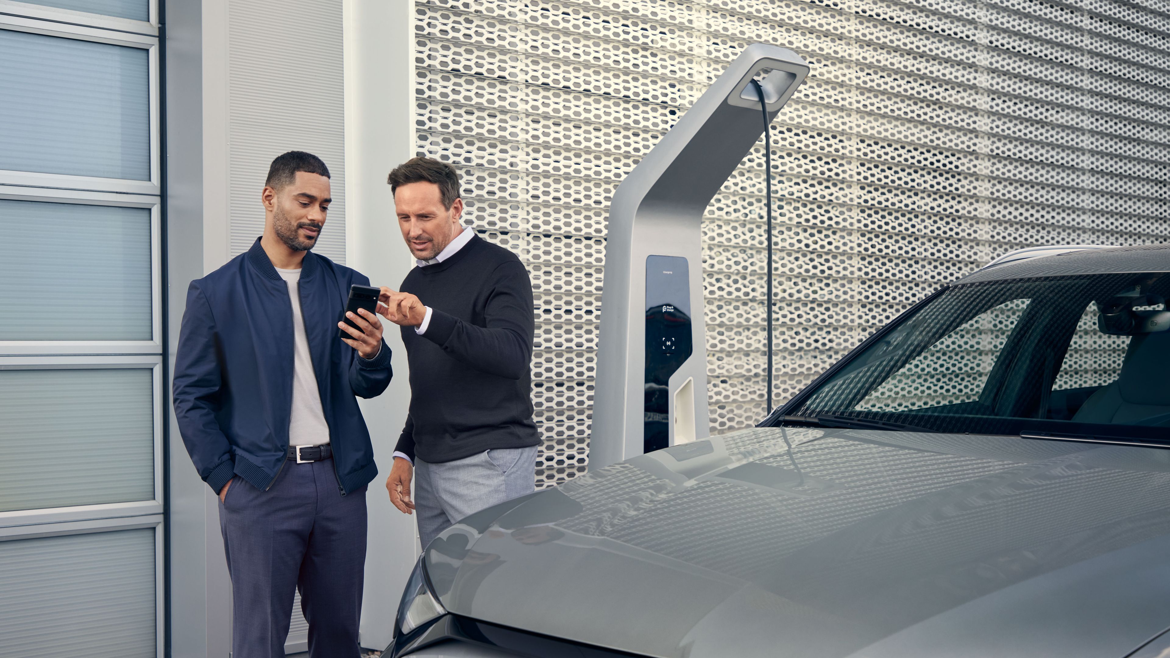 Ein Audi Service Mitarbeiter und ein Kunde blicken auf ein Smartphone, im Vordergrund steht ein Audi Elektroauto